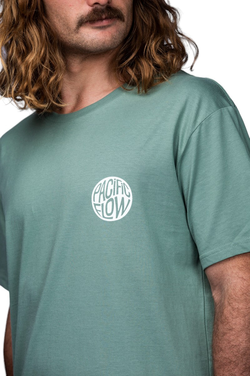 A sage coloured shirt with an island on the back, with surfboards, a fire, palmtrees, a tent, and waves to surf.